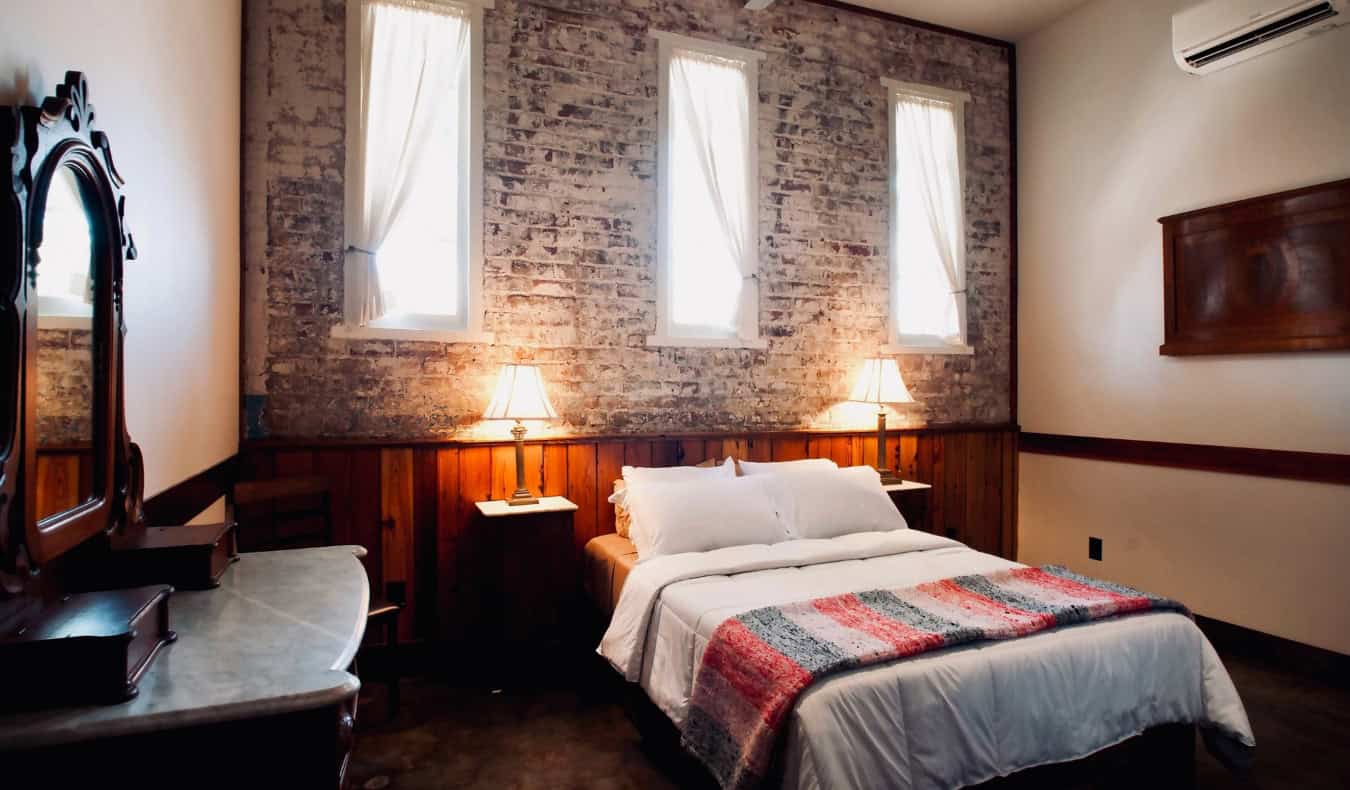 Tempat tidur ukuran queen di kamar tamu dengan dinding bata terbuka dan perabotan antik di The Inn at the Old Jail, sebuah hotel di New Orleans, Louisiana